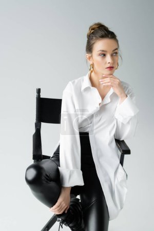 Foto de Fashionable woman in white shirt and black latex pants sitting on chair and looking away isolated on grey - Imagen libre de derechos