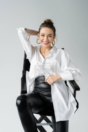 Foto de Cheerful woman in white shirt and black latex pants smiling at camera while posing on chair isolated on grey - Imagen libre de derechos