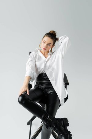 Foto de Stylish woman in black tight pants and white shirt holding hand behind head while sitting on chair on grey background - Imagen libre de derechos