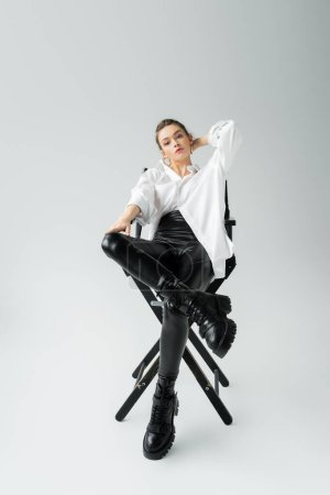 volle Länge der trendigen Frau in schwarzen Latex-Hosen und groben Stiefeln posiert auf Stuhl auf grauem Hintergrund