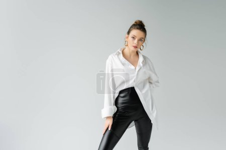 Foto de Stylish woman in black tight pants and white oversize shirt posing and looking at camera isolated on grey - Imagen libre de derechos