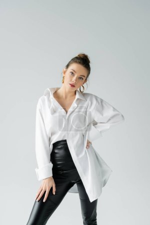 Foto de Fashionable woman in black tight pants and white oversize shirt posing with hand on hip isolated on grey - Imagen libre de derechos