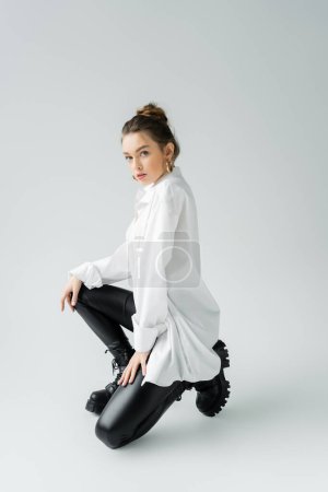 Foto de Full length of young woman in white shirt and black rough boots looking at camera while posing on grey background - Imagen libre de derechos