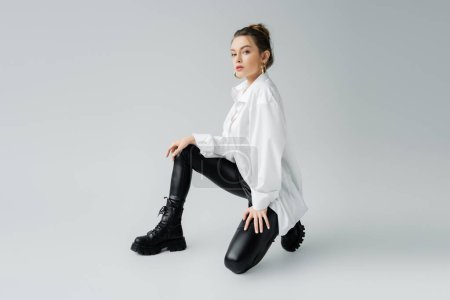 Photo for Full length of trendy model in black latex pants and oversize shirt looking at camera on grey background - Royalty Free Image