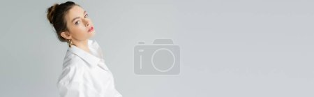 portrait of trendy woman with natural makeup posing in white oversize shirt isolated on grey, banner