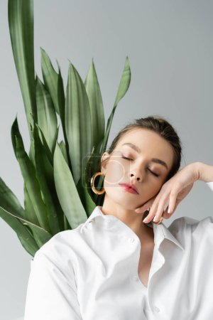 portrait of sensual woman holding hand near face while posing with closed eyes near green plant isolated on grey