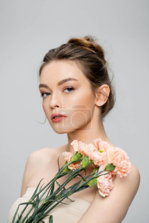 Photo for Pretty young woman with bare shoulders looking at camera near bouquet of carnations isolated on grey - Royalty Free Image