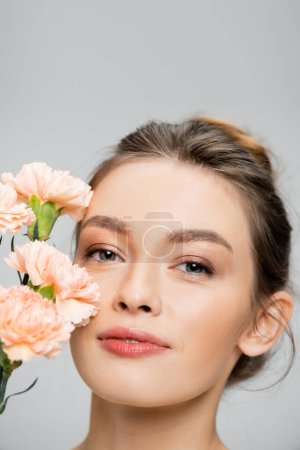 Photo for Portrait of charming woman with natural makeup on perfect face looking at camera near carnations isolated on grey - Royalty Free Image