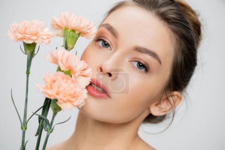 Photo for Young woman with perfect skin and natural makeup looking at camera near fresh carnations isolated on grey - Royalty Free Image