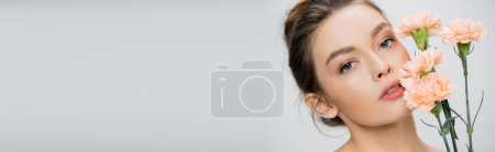 junge und hübsche Frau mit natürlichem Make-up, die in die Kamera schaut, in der Nähe frischer Nelken isoliert auf grau, Banner