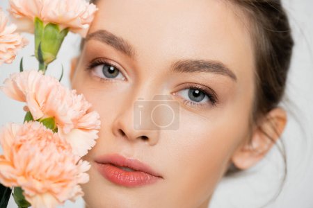 Photo for Close up portrait of pretty woman with clean skin and natural makeup near peach carnations isolated on grey - Royalty Free Image
