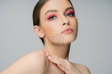 Téléchargez les photos : Young woman with perfect skin and makeup touching bare shoulder and looking at camera isolated on grey - en image libre de droit