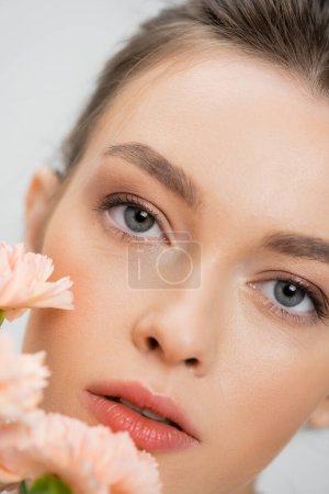 close up portrait of young woman with natural makeup looking at camera near blurred flowers isolated on grey