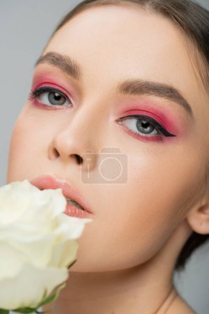 close up portrait of woman with pink eye shadows looking at camera near ivory rose isolated on grey