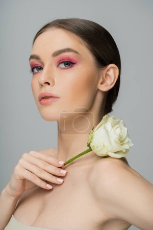 sensual woman with pink makeup holding fresh rose near naked shoulder and looking at camera isolated on grey