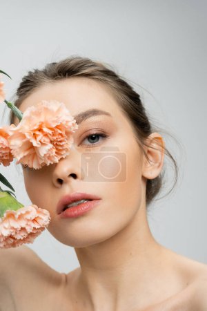 junge Frau mit perfekter Haut und natürlichem Make-up verschleiert Gesicht mit Pfirsichnelken isoliert auf grau