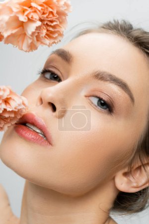 Photo for Close up of charming woman with perfect face and natural makeup looking at camera near flowers isolated on grey - Royalty Free Image