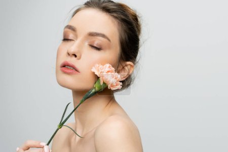 young woman with naked shoulders and natural makeup posing with fresh carnation isolated on grey