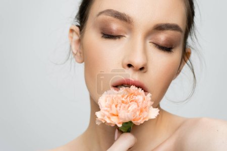 young woman with makeup and closed eyes holding peach carnation near face isolated on grey