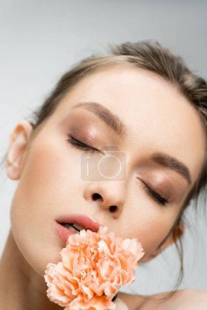 close up portrait of young woman with makeup and closed eyes posing with carnation isolated on grey