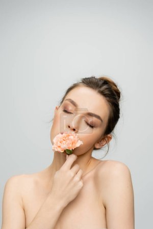 young woman with closed eyes and makeup smelling aromatic carnation isolated on grey