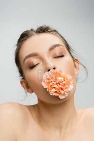 junge Frau mit geschlossenen Augen posiert mit Pfirsichnelke im Mund isoliert auf grau