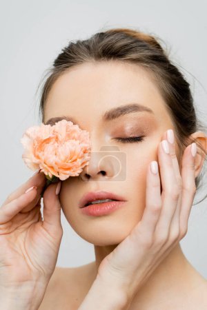 mujer sensual con piel perfecta tocando la cara y cubriendo el ojo con clavel de melocotón aislado en gris