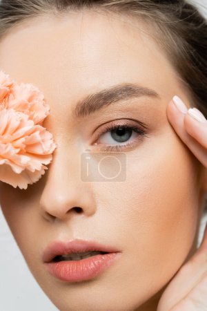 primer plano retrato de la mujer con la cara perfecta y maquillaje natural que cubre el ojo con clavel de melocotón aislado en gris