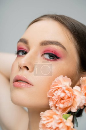 peach carnations near sensual woman with pink makeup looking at camera isolated on grey