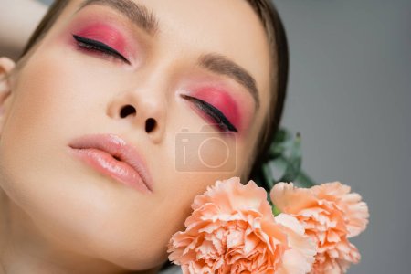 Téléchargez les photos : Close up portrait of young woman with pink makeup posing with closed eyes near carnations isolated on grey - en image libre de droit