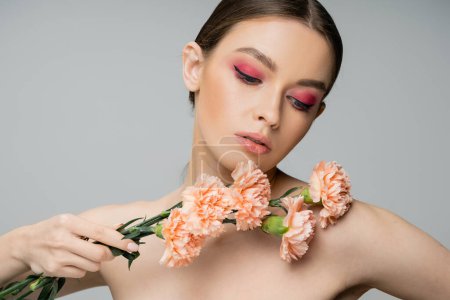 Photo for Pretty woman with pink visage holding fresh carnations near bare shoulder isolated on grey - Royalty Free Image