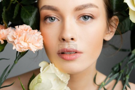 portrait of young woman with natural makeup and perfect skin looking at camera near fresh flowers isolated on grey