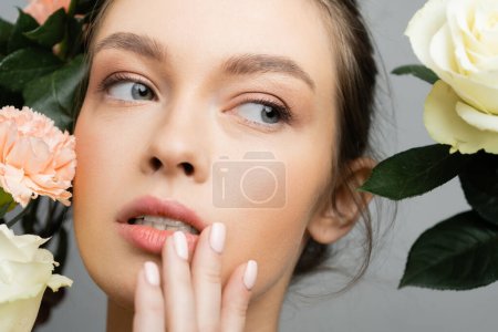 Foto de Encantadora mujer con cara perfecta y maquillaje natural tocando los labios cerca de flores frescas aisladas en gris - Imagen libre de derechos