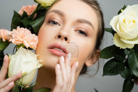 portrait of sensual woman touching face while looking at camera near roses and carnations isolated on grey
