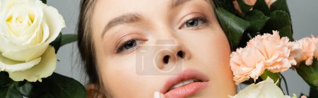 close up portrait of pretty woman with natural makeup looking at camera near flowers isolated on grey, banner