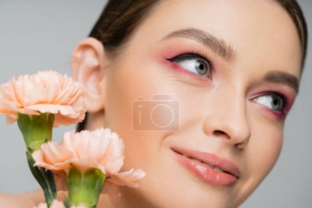 Photo for Close up portrait of smiling woman with makeup looking away near peach carnations isolated on grey - Royalty Free Image