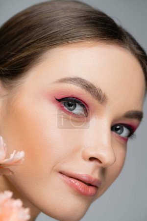 close up portrait of young woman with pink makeup looking at camera near floral petals isolated on grey