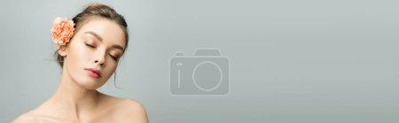 Foto de Encantadora mujer con hombros desnudos y flor de clavel en el pelo posando con los ojos cerrados aislados en gris, bandera - Imagen libre de derechos