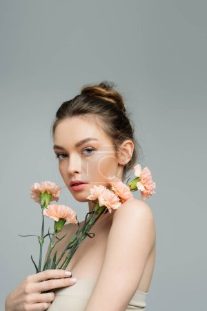Photo for Pretty young woman with natural makeup and bare shoulders looking at camera near peach carnations isolated on grey - Royalty Free Image