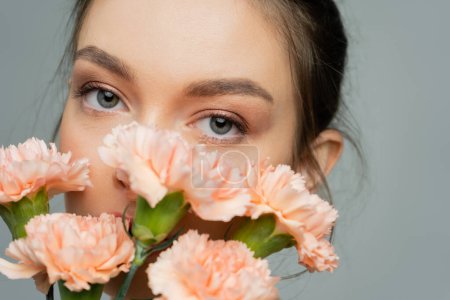 portrait of young woman looking at camera near peach carnations isolated on grey