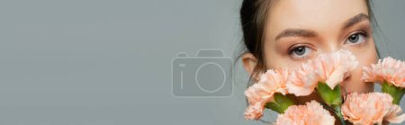 Photo for Young woman looking at camera behind fresh carnations isolated on grey, banner - Royalty Free Image
