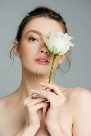 young woman with naked shoulders and perfect skin obscuring face with white rose isolated on grey