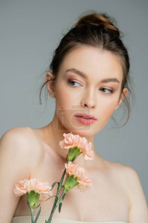 Photo for Charming woman with naked shoulders and perfect skin posing with peach carnations isolated on grey - Royalty Free Image