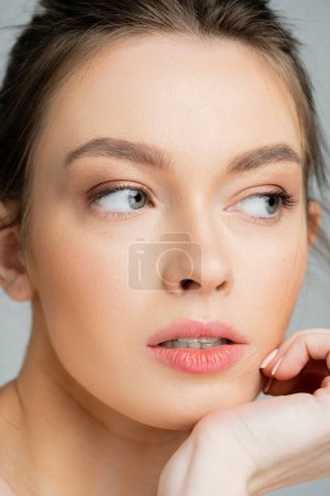 close up portrait of young woman with perfect face and natural makeup holding hand near chin isolated on grey