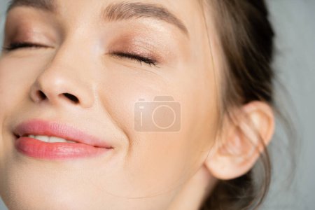 Close up view of pleased young woman with natural makeup closing eyes isolated on grey 
