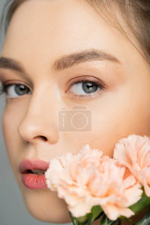 Close up view of young model looking at camera near carnations isolated on grey 