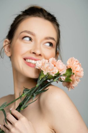 Cheerful young woman with naked shoulders holding carnations isolated on grey 
