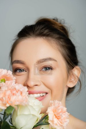 Cheerful young woman looking at camera near blurred bouquet isolated on grey 