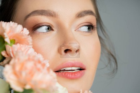 Close up view of woman with natural visage looking away near blurred carnations isolated on grey 
