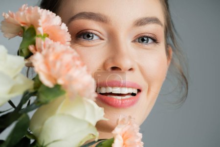 Smiling fair haired woman looking at camera near blurred roses and carnations isolated on grey 